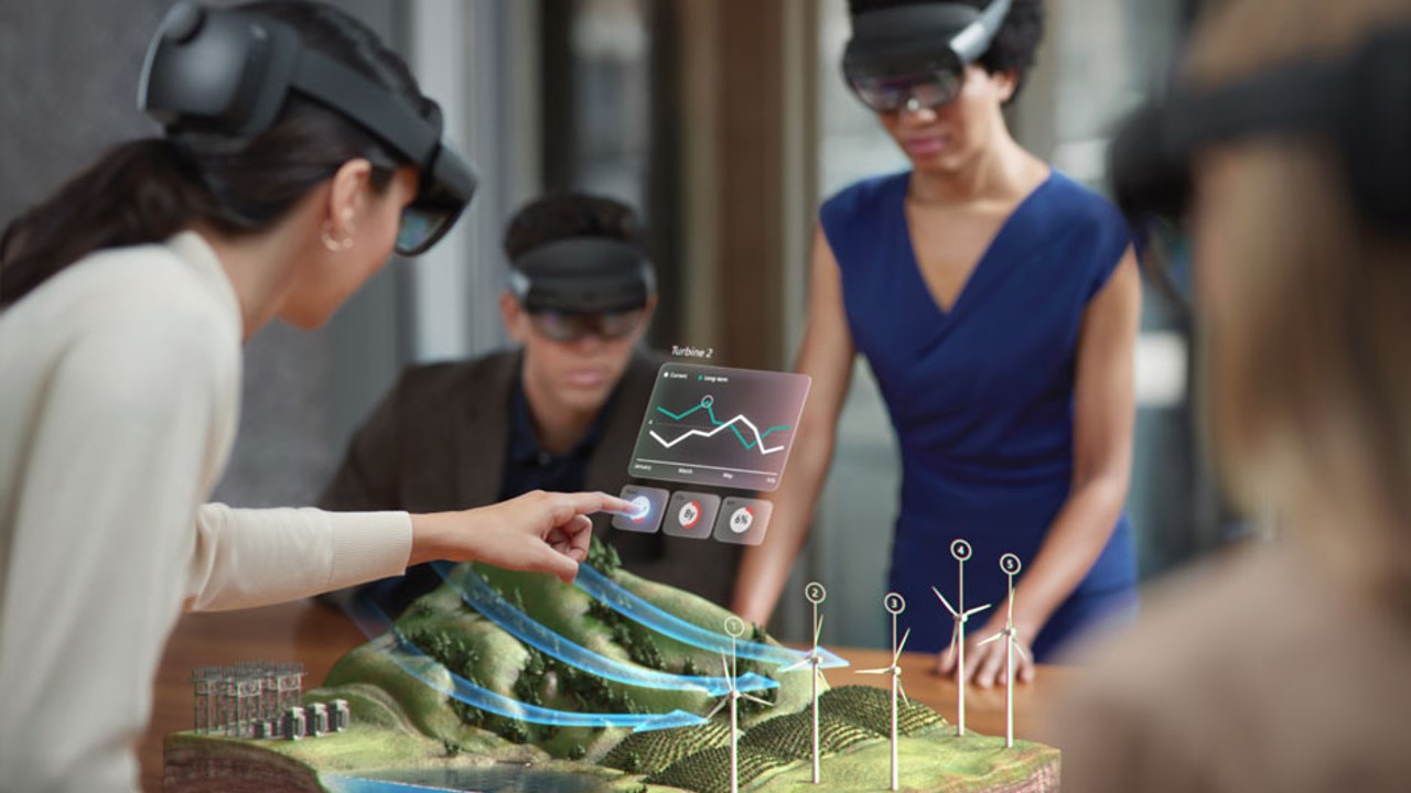 Photo of multiple users wearing HoloLens devices and interacting with an immersive experience on a table showing terrain with wind turbines.