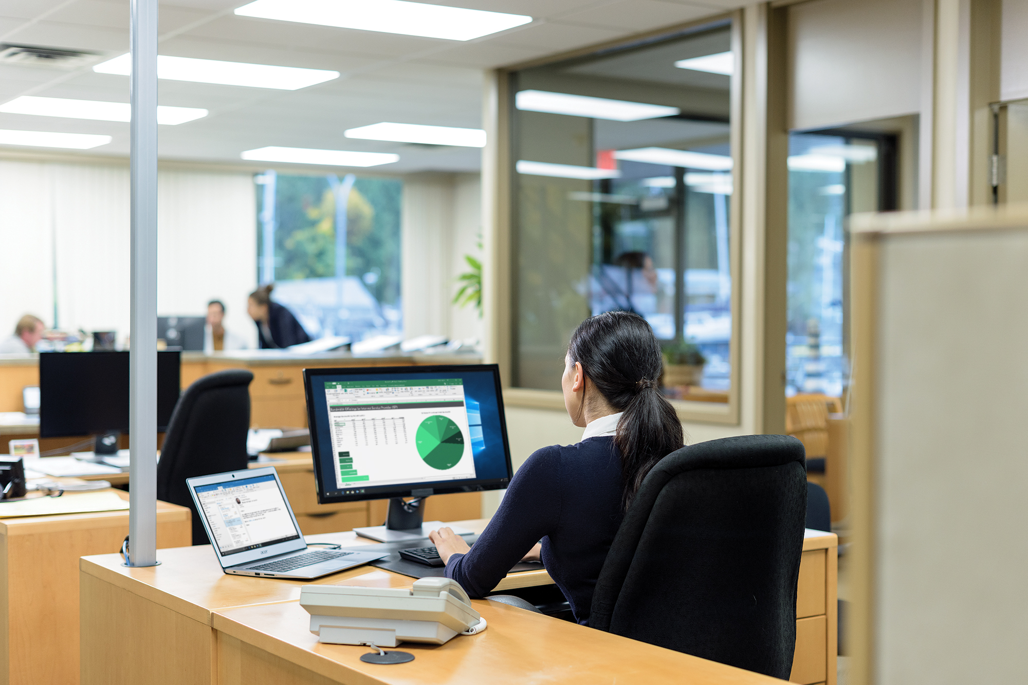 En kontorsarbetare tittar på diagram och tabeller på en skärm medan andra träffas i bakgrunden.
